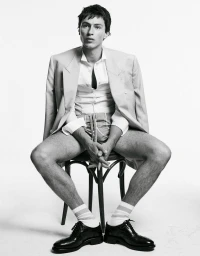 Man seated on a stool wearing a blazer draped over his shoulders, white shirt, and patterned shorts against a white backdrop.