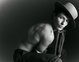 Black and white portrait of a shirtless male model wearing a hat, seated backwards on a chair for Knack Weekend.