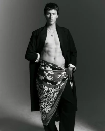 Black and white portrait of a male model wearing an open dark coat and patterned wrap against a grey studio backdrop.