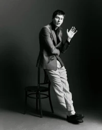 Man wearing a blazer and trousers leaning against a wooden chair in a studio portrait for Knack Weekend
