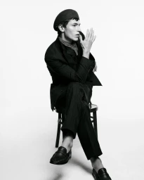 Monochrome fashion portrait of male model Clément Laguardia seated on a chair wearing a dark suit and beret against white seamless.