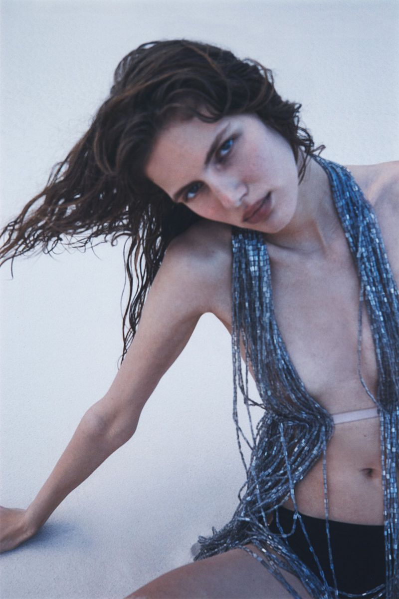 Brunette woman with beaded top poses against a light background in a fashion shot. Her hair is loose and slightly tousled.