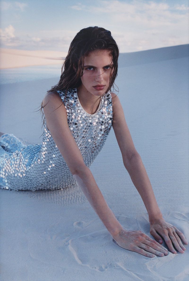 A slender brunette woman is lying on the light-colored sand in a sequined dress. The photo is taken outdoors on a sunny day.