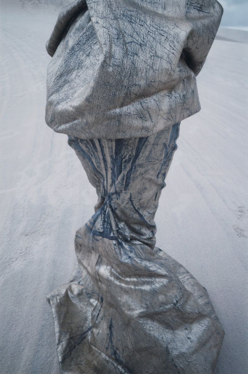 Abstract draped fabric sculpture with a cracked texture, casting shadows on a grainy surface, creating a surreal, otherworldly visual.