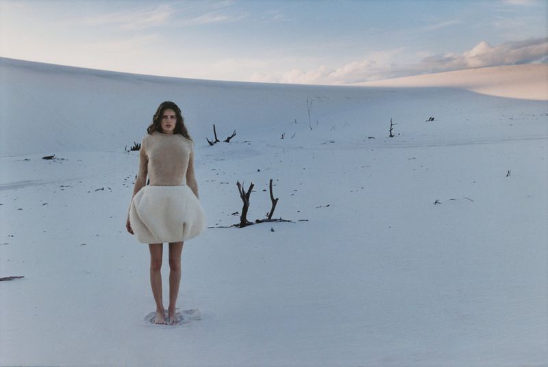 A woman stands barefoot in the snow wearing a beige top and a puffy white skirt with a snow covered landscape background with dead trees.