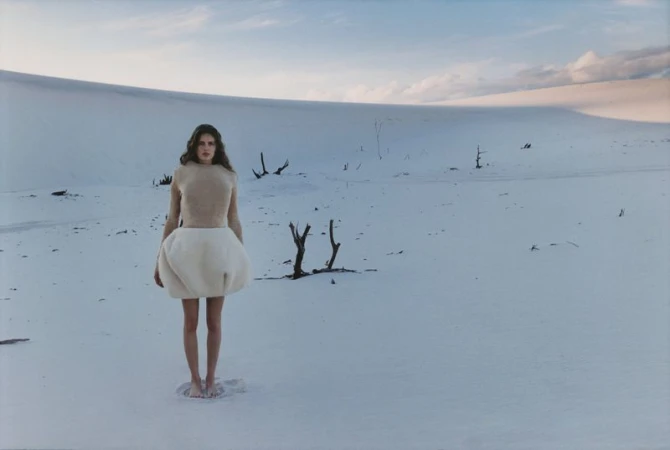 A woman stands barefoot in the snow wearing a beige top and a puffy white skirt with a snow covered landscape background with dead trees.