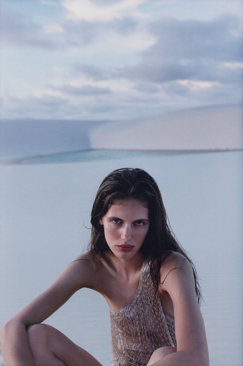 Portrait of a woman in a gold, shimmering top, seated in front of light blue water and white sand dunes under a cloudy sky.