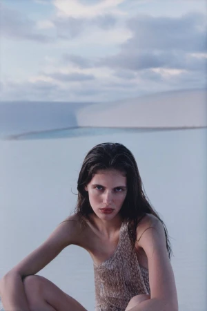 Portrait of a woman in a gold, shimmering top, seated in front of light blue water and white sand dunes under a cloudy sky.