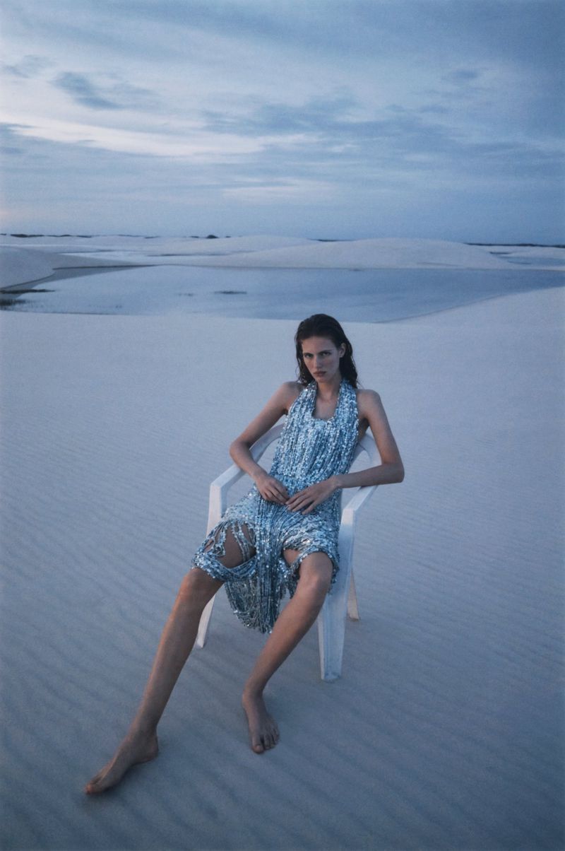 A woman in a shimmering halter dress sits on a white chair in a pale desert landscape under a cloudy sky in an outdoor fashion shot.