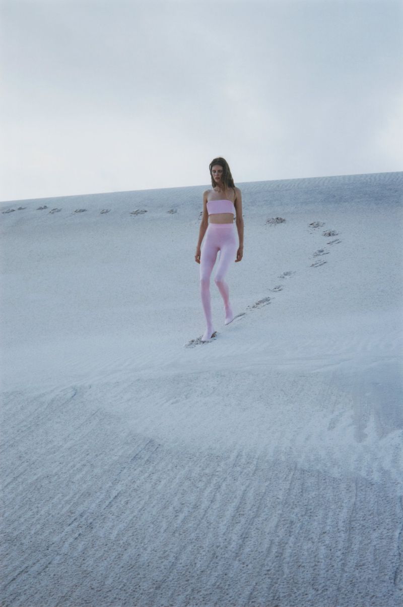 A slender woman in light pink athleticwear walks across a white sand dune under a cloudy sky, leaving a trail of footprints.
