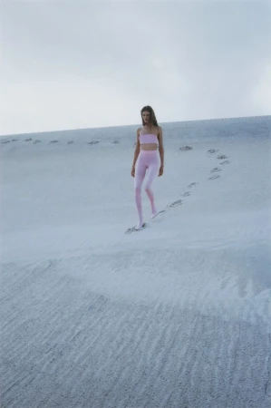 A slender woman in light pink athleticwear walks across a white sand dune under a cloudy sky, leaving a trail of footprints.