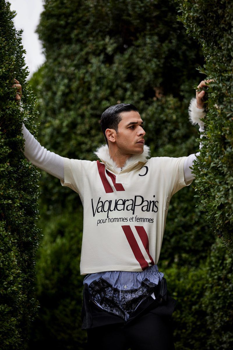 Adult male poses in a fashion shot outdoors, wearing a cream-colored shirt with dark red stripes and the text 'Vaquera Paris'.