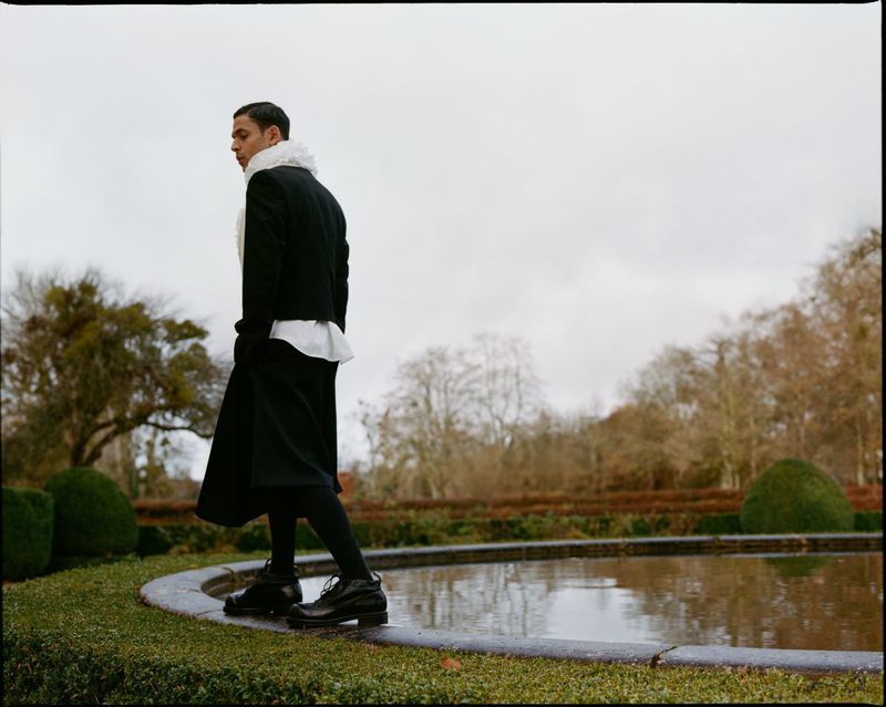 Man walking in black coat and skirt near a reflecting pool in a garden setting on a cloudy day, fashion editorial photography.
