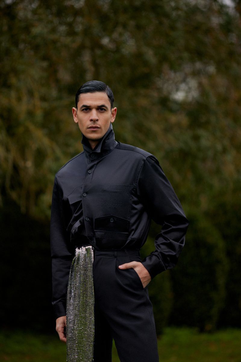 Portrait of a man wearing a black button-up shirt and pants with silver fabric detail posing outdoors in front of green foliage.