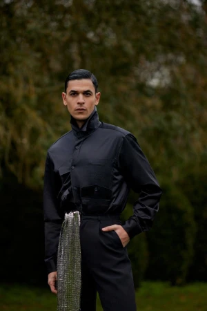 Man wearing a black shirt and trousers with a metallic embellished sash standing outdoors in a park.