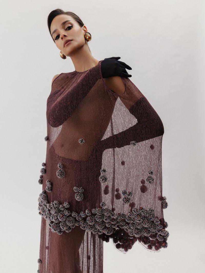 A woman wearing a translucent, embellished dress with long gloves poses against a white backdrop in a studio, showcasing high-fashion elegance.