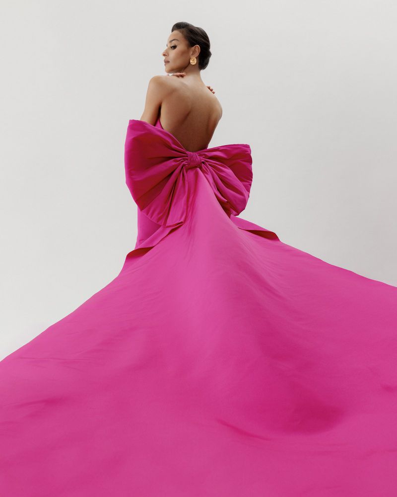 Brunette woman in a bright pink, strapless gown with a large bow, standing in a photo studio with an off-white backdrop. Fashion shot, back view.