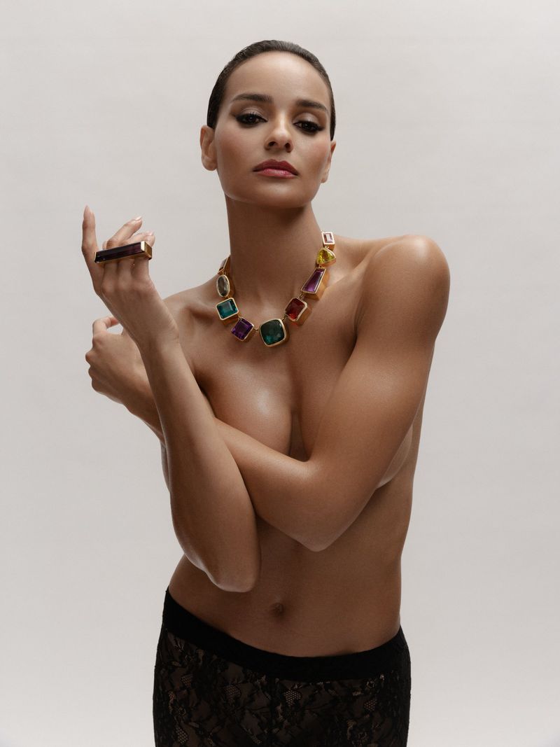 Attractive brunette woman poses in studio, wearing a colorful gemstone necklace and ring. Bright lighting emphasizes her skin and jewelry.