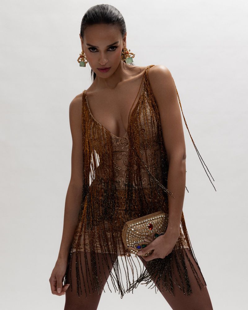 Elegant woman in a short brown beaded dress and statement earrings holds a decorative gold clutch in a bright studio shot.
