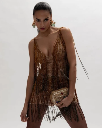 Model Gracie Carvalho wearing a beaded fringe dress and ornate earrings against a white studio background.