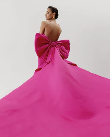 Model Gracie Carvalho wearing a dramatic magenta gown with a large bow and long train against a white background