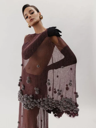 Model wearing a sheer brown textured dress with black gloves and round earrings against a plain white studio background.