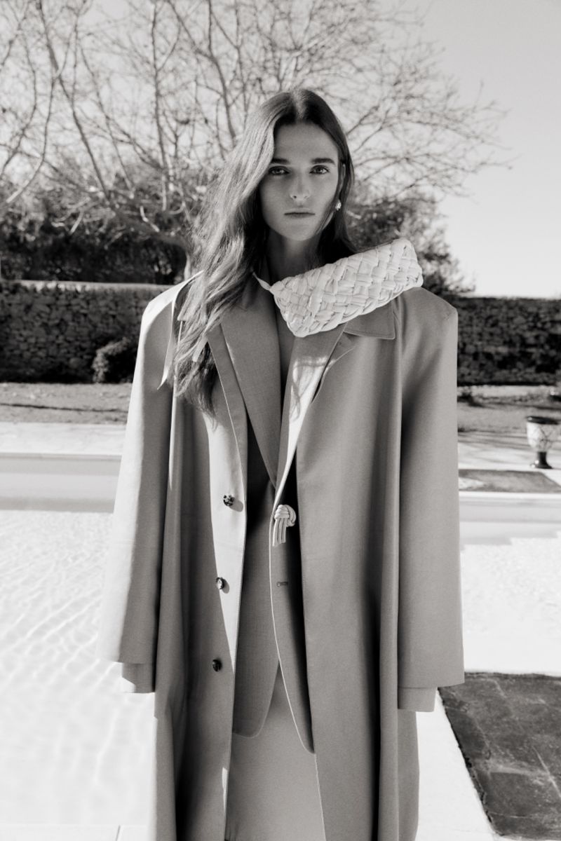 A young woman in a beige coat and matching suit poses outdoors with a small woven handbag in a monochromatic fashion shot.