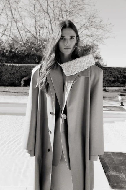 A young woman in a beige coat and matching suit poses outdoors with a small woven handbag in a monochromatic fashion shot.