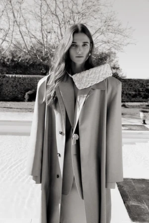 A young woman in a beige coat and matching suit poses outdoors with a small woven handbag in a monochromatic fashion shot.