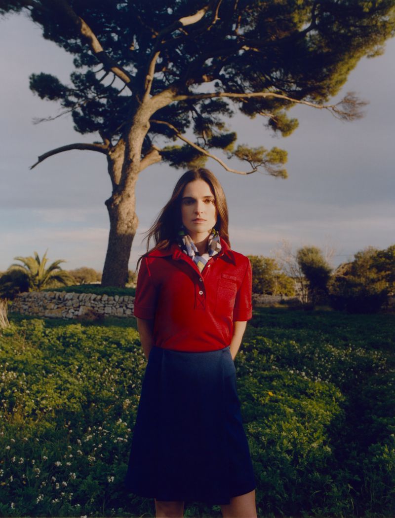 An adult woman stands outside in a red shirt and blue skirt near a large tree with green grass and a cloudy sky in the background.