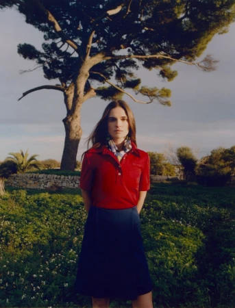 An adult woman stands outside in a red shirt and blue skirt near a large tree with green grass and a cloudy sky in the background.