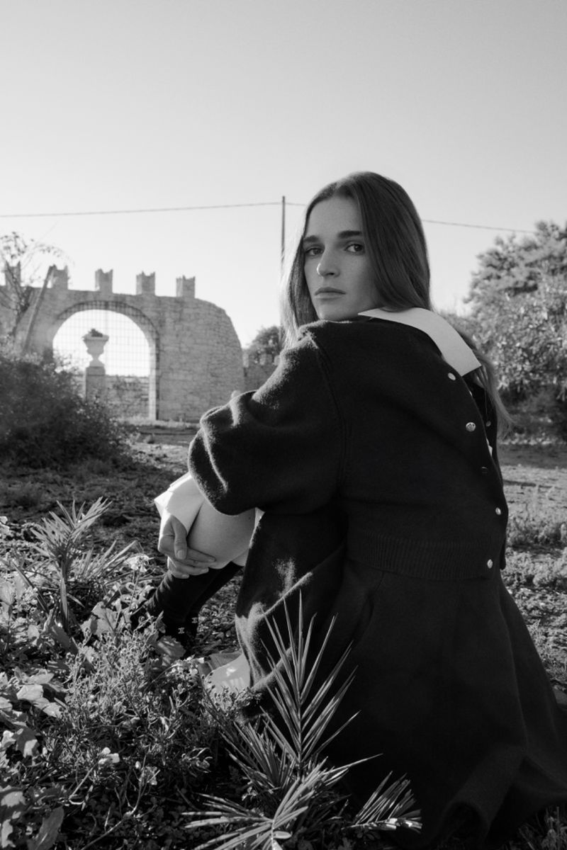 Young adult woman poses in nature, wearing a dark coat with a white collar, sitting near stone archway with greenery. Black and white.