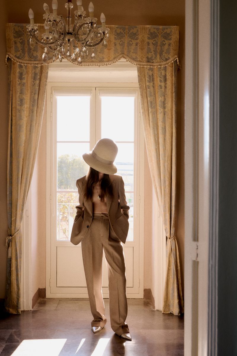 Woman in beige pantsuit and wide-brimmed hat poses in sunlit hallway with chandelier and patterned curtains at large window.