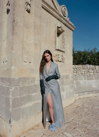 Model leaning against an ornate stone wall wearing a long grey coat, dark gloves, and fuzzy shoes.