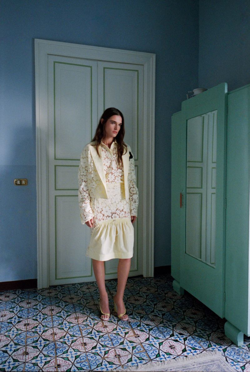 A woman in a light yellow lace dress and jacket stands in a room with patterned tile flooring and a light blue wall with white trim.