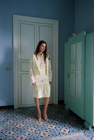 A woman in a light yellow lace dress and jacket stands in a room with patterned tile flooring and a light blue wall with white trim.