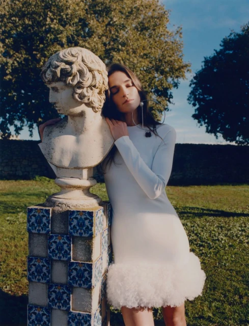 A young adult woman in a white dress poses beside a classical bust outdoors, with soft sunlight highlighting the scene's textures and colors.