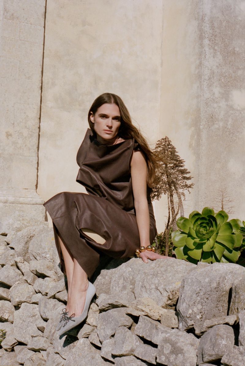 An adult female model sits on a stone wall wearing a brown dress and light grey and black shoes in an outdoor setting.