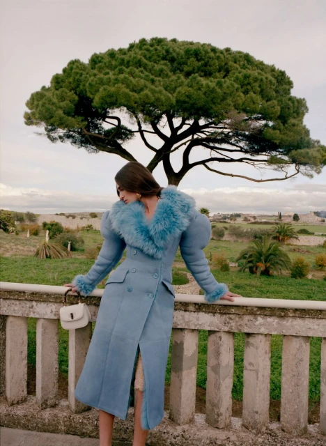 A woman in a light blue coat with a fur collar and cuffs leans against a stone railing in front of a large green tree.