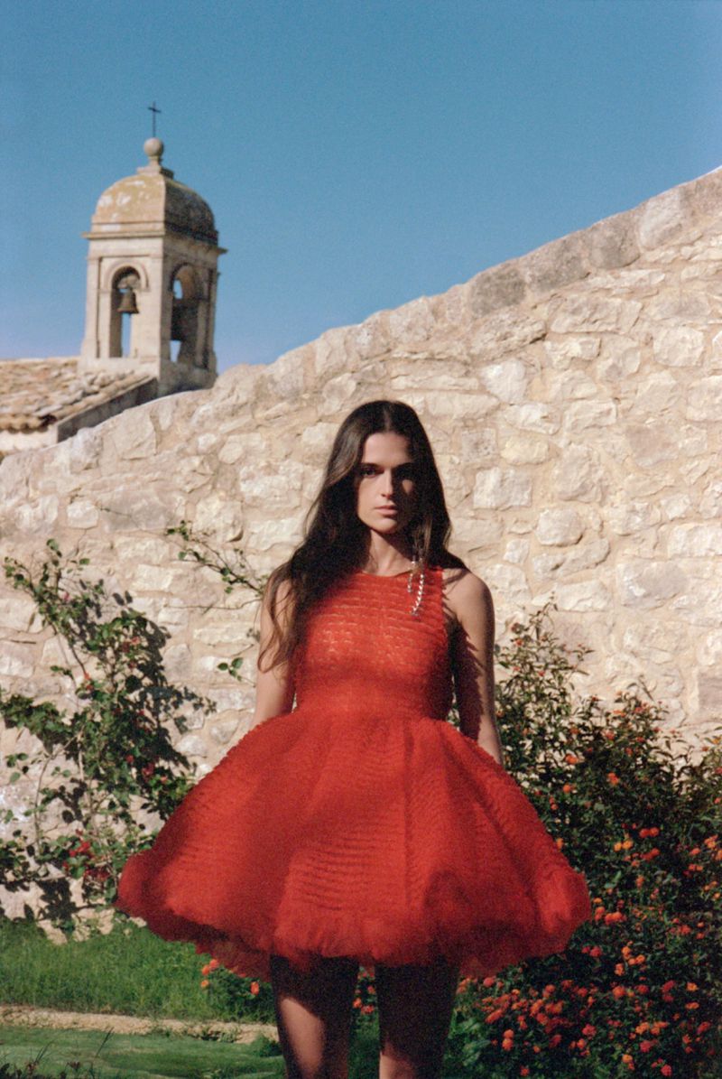 A woman poses in a red dress with a stone building and tower in the background under a clear blue sky, creating a fashion editorial shot.