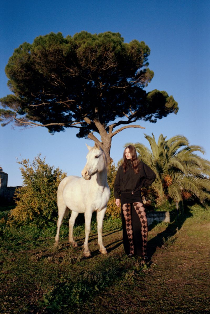 A woman standing next to a white horse in a field with a large tree in the background under a clear blue sky during daytime.