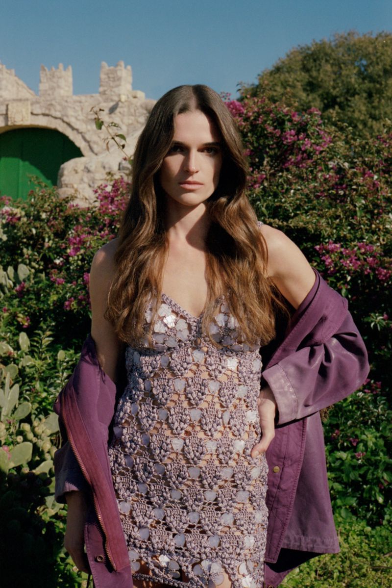 Model wearing a lavender crocheted dress and a purple jacket standing outdoors before a stone structure and flowering bushes.