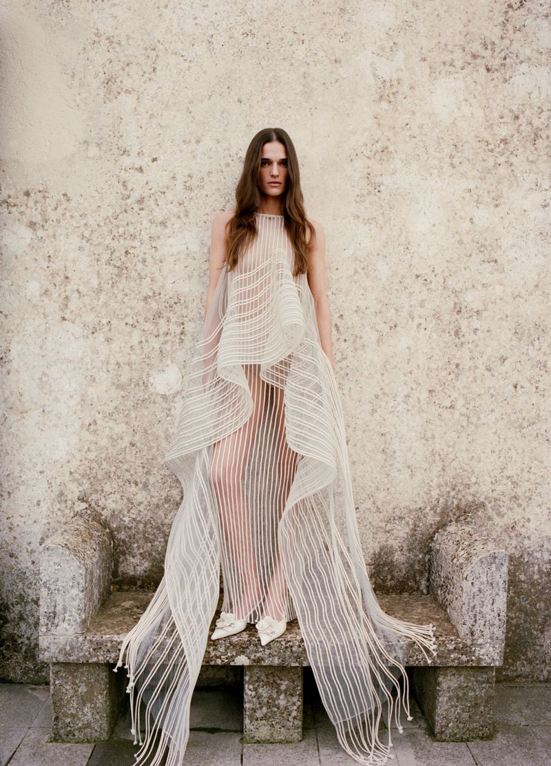 A woman in a flowing white dress stands on a stone bench against a textured wall, exuding a minimalist fashion aesthetic with long hair.