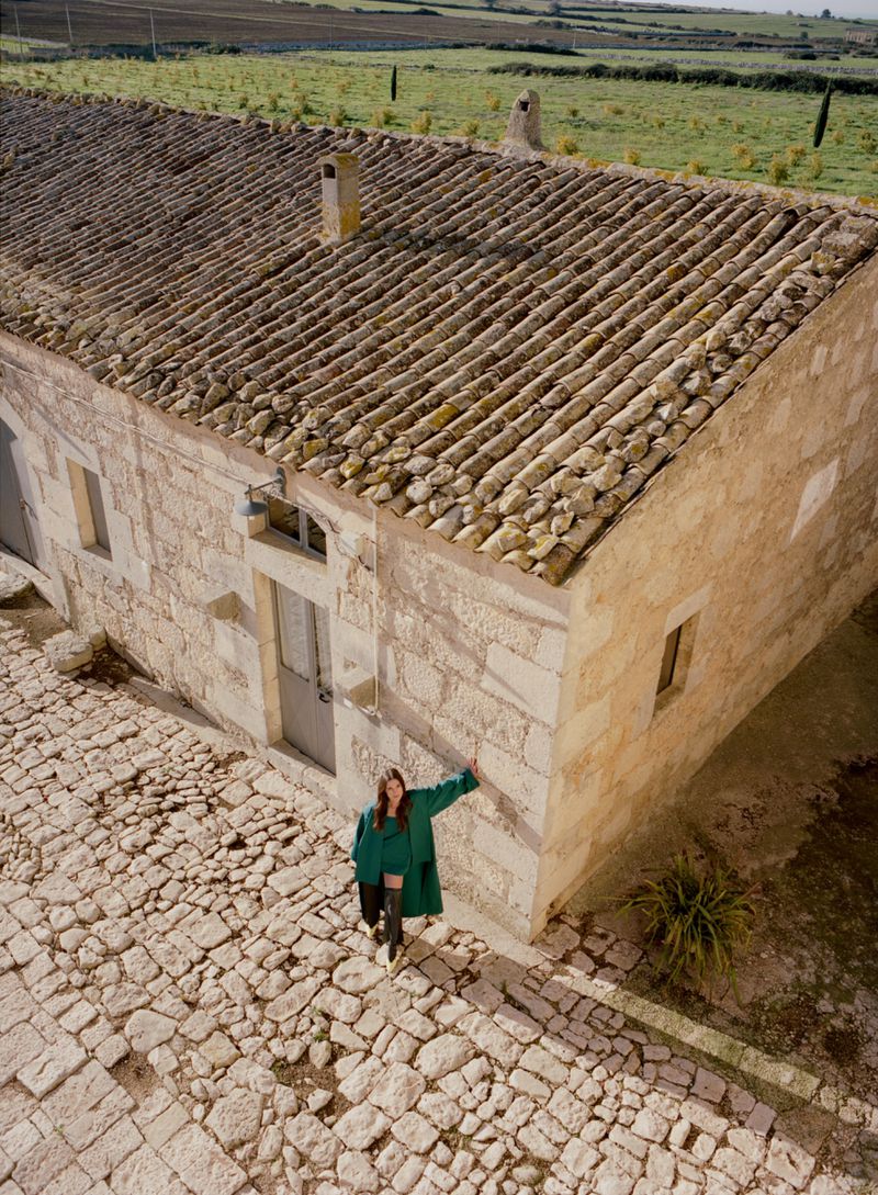 Woman in a green coat stands on a cobblestone street next to an old stone building with a tile roof in a rural setting.