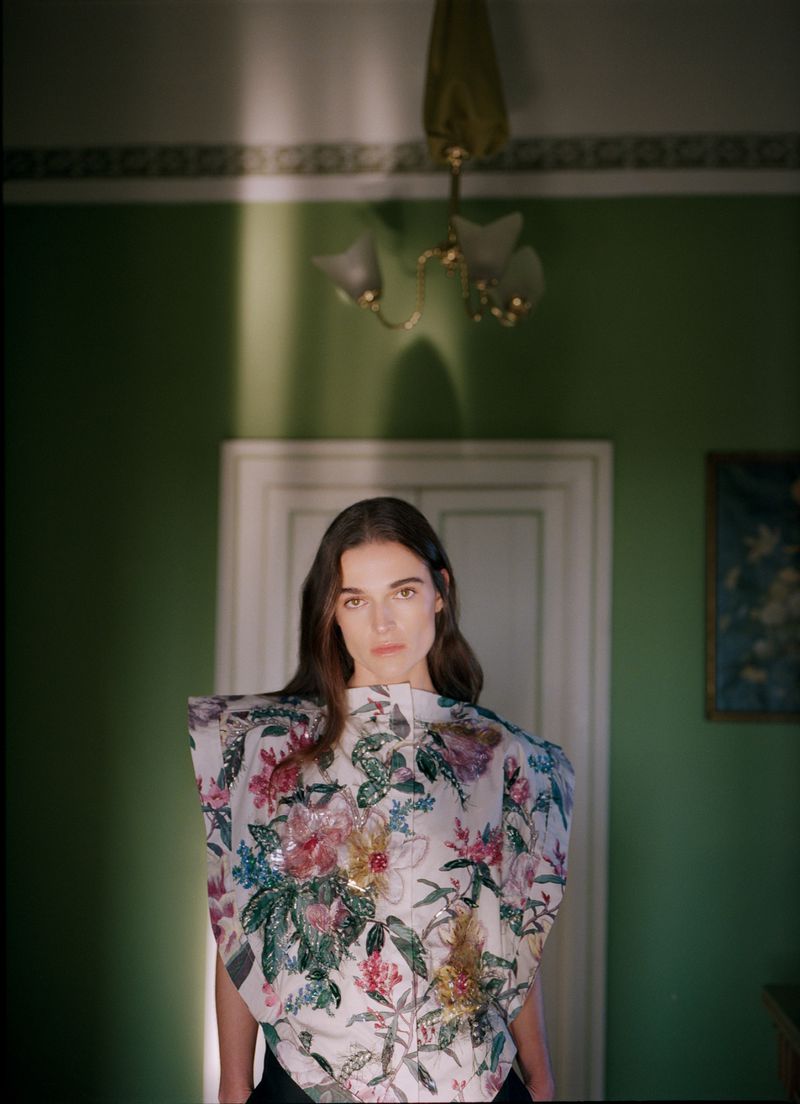 Young woman in a floral top stands indoors against a green wall with doorway, a chandelier hangs above her in a fashion photography shot.