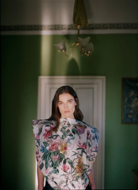 Young woman in a floral top stands indoors against a green wall with doorway, a chandelier hangs above her in a fashion photography shot.