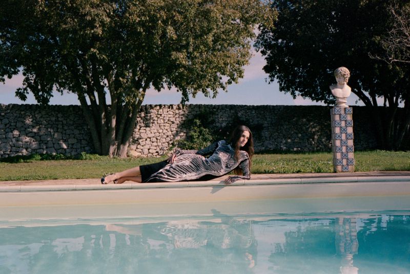 An adult female poses by a swimming pool in a gray patterned dress, with stone walls, trees, and a bust statue in the background.