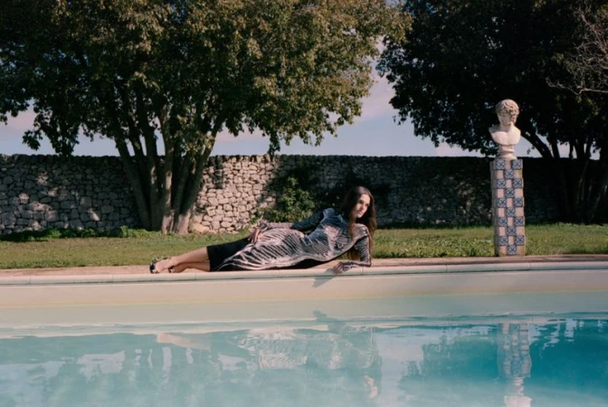 An adult female poses by a swimming pool in a gray patterned dress, with stone walls, trees, and a bust statue in the background.