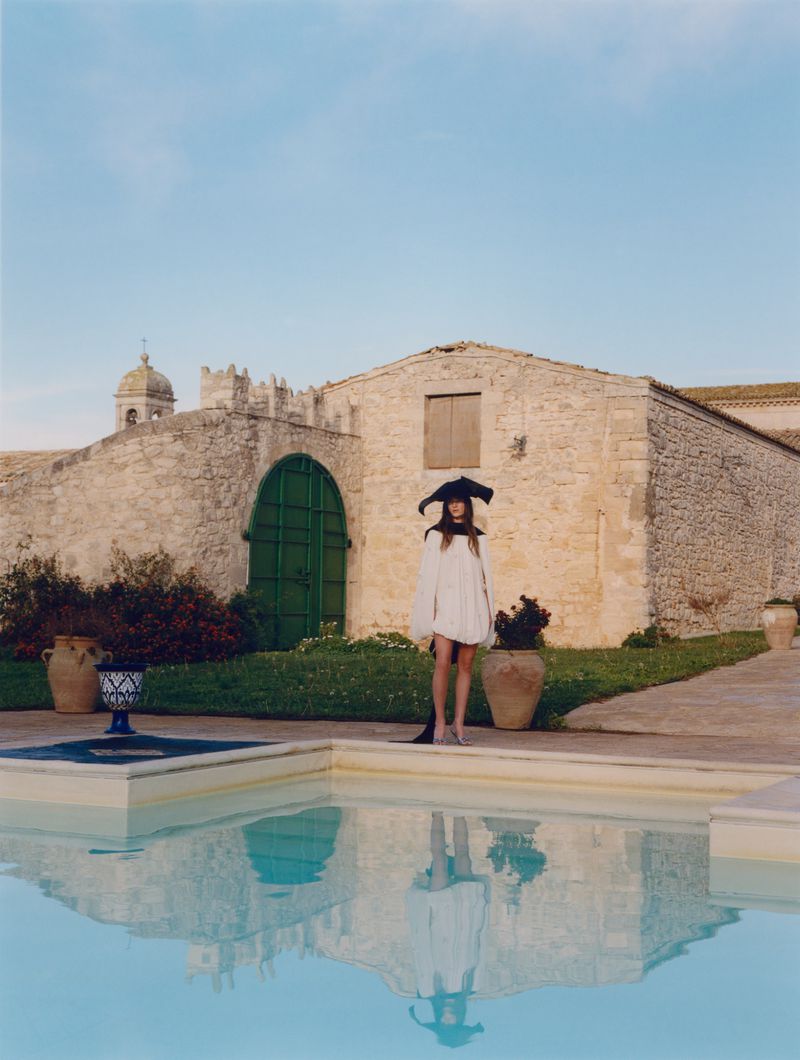 A slender woman in a white dress and distinctive black hat stands near a pool at an outdoor stone villa on a clear day.
