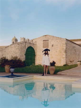 A slender woman in a white dress and distinctive black hat stands near a pool at an outdoor stone villa on a clear day.